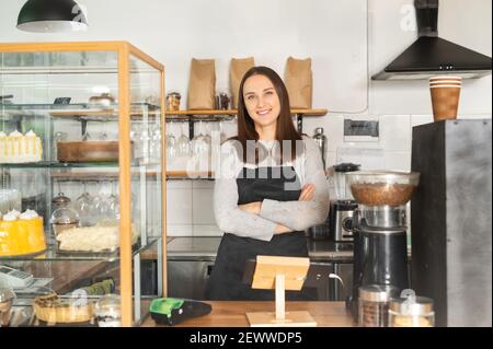 Selbstbewusste Geschäftsinhaberin, die mit gekreuzten Armen an der Theke eines Restaurants steht, blickt die Backwarenfrau im Vorfeld auf die Kamera und lächelt Stockfoto