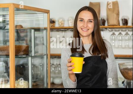 Freundliche und lächelnde Kellnerin im Schürze hält Papiertasse mit Take-Away-Kaffee in die Kamera. Fröhliche weibliche Bäckereibesitzerin bereitete heiße Getränke zu Stockfoto