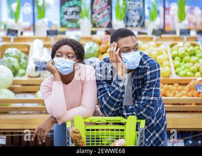 Gelangweilte Millennial Paar in Gesichtsmasken auf Warenkorb gelehnt, krank und müde von der Herstellung von Einkäufen in der Mall Stockfoto