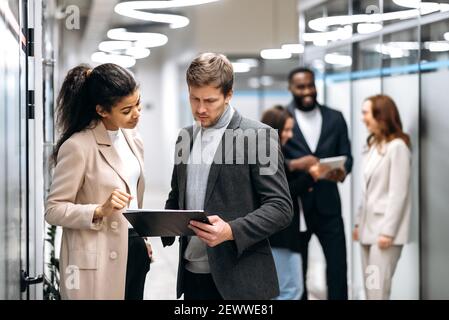 Seriöse multirassische Kollegen stehen in der Bürohalle, reden über Arbeitsfragen. Konzentrierte Geschäftspartner diskutieren Unternehmensprojekt, geben sich gegenseitig Ratschläge, Teamwork-Konzept Stockfoto