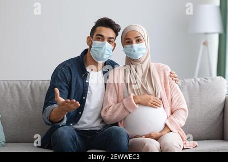 Bleiben Sie Zu Hause. Schwangere Muslimische Paar In Medizinischen Masken Sitzen Auf Couch Stockfoto