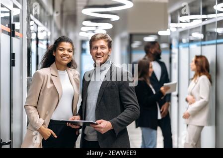 Porträt von zwei multirassischen zufriedenen erfolgreichen Kollegen in formeller Kleidung, die in einem modernen Büro stehen und freundlich lächeln auf die Kamera Stockfoto