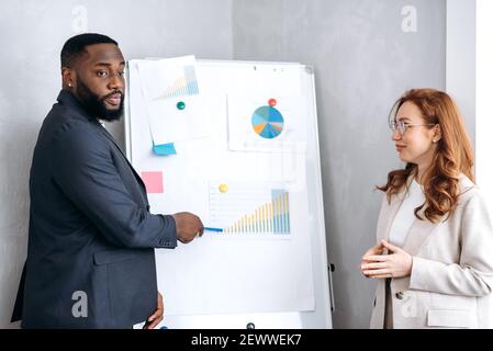Afroamerikanischer Mann in stilvollem Anzug Präsentation Arbeit Bericht auf Konferenz mit lächelnden kaukasischen Dame. Einflussreiche Geschäftspartner, die mit Finanzgrafiken in der Nähe des Flipchart stehen und darüber sprechen Stockfoto