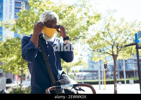 afroamerikanischer älterer Mann mit Gesichtsmaske auf dem Fahrrad sitzend Auf der Straße Ohrhörer aufsetzen Stockfoto