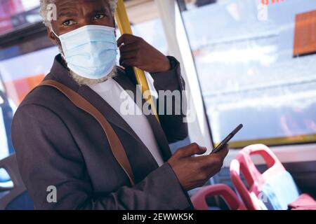 afroamerikanischer älterer Mann mit Gesichtsmaske auf Bus stehend Mit dem Smartphone Stockfoto