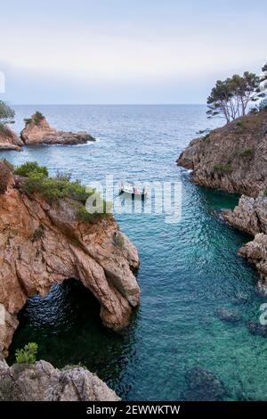 Eine kleine Bucht mit kristallklarem Wasser und mit einer kleinen Höhle, segelt ein kleines Boot mit drei Personen, Cala de Sa Foradada, Palamos, Costa Brava Stockfoto