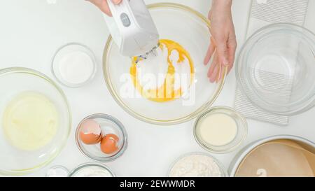 Frau Hände mischen Eigelb mit Zucker in einer Glasschüssel mit einem elektrischen Mixer. Nahaufnahme Backvorgang, flach legen Stockfoto