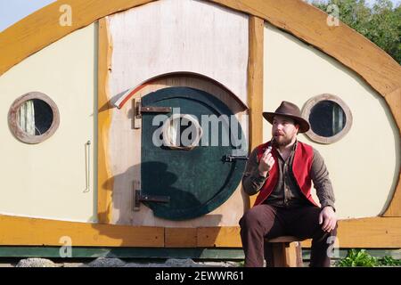 Junger Mann in einer roten Weste mit Pfeife und Hut von einem Hobbit Hole Hut, Camping in Cornwall, UK. Stockfoto