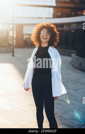 Lockig behaarte Geschäftsfrau mit Brille posiert draußen und Lächeln Sie an einem sonnigen Tag auf die Kamera Stockfoto