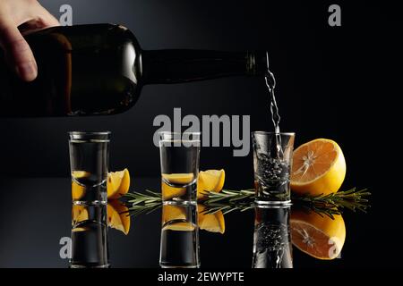 Gin wird in ein kleines Glas aus einer antiken Flasche aus dunklem Glas gegossen. Ein starkes alkoholisches Getränk mit Zitrone und Rosmarin auf einem schwarzen reflektierenden Hintergrund Stockfoto