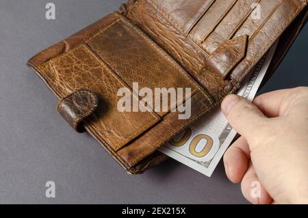 Eine Hand zieht einen Dollarschein aus einer braunen Lederbörse auf grauem Hintergrund heraus. Einhundert amerikanische Dollar-Rechnung. Erwachsener Mann. Selektiver Fokus. Stockfoto