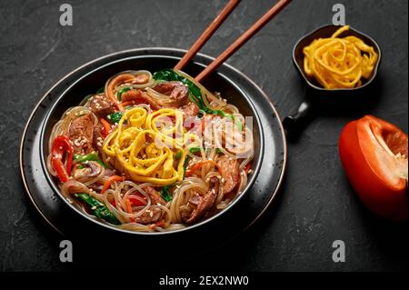Japchae in schwarzer Schale auf dunkler Schieferplatte. Koreanische Küche Glas Chapchae Nudeln Gericht mit Gemüse und Fleisch. Traditionelle asiatische Küche. Authentisch Stockfoto