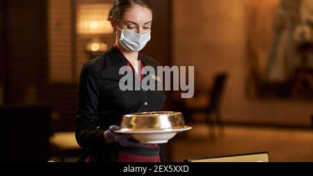 Elegante junge Frau in Uniform und Gesichtsmaske trägt ein Teller mit Deckel beim Servieren von Tischen im Restaurant Stockfoto
