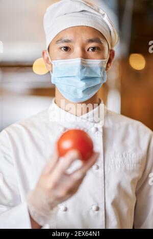 Portrait Foto von jungen asiatischen Koch in schützende Gesichtsmaske Handschuhe tragen und Tomate halten Stockfoto