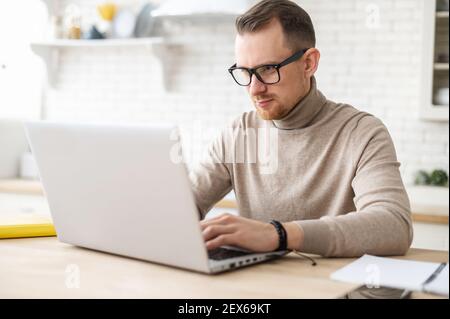 Serious fokussierten bestimmten jungen bärtigen Geschäftsmann oder Freiberufler in Brille und Freizeitkleidung sitzen am Küchentisch, tippen auf Laptop, arbeiten an Projekt, Recherche, E-Mail an Kunden schreiben Stockfoto