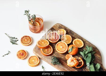 Ganze und in Scheiben geschnittene Mandarinen und Blutorangen mit Thymian, Minzkräutern auf Holzbrett. Weiße Tischhintergründe. Limonade trinken Stockfoto