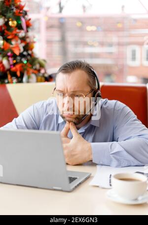 Ein müder Kaukasusmann mittleren Alters mit einem Headset in einer Brille, das auf den Laptop-Bildschirm fokussiert ist. Er arbeitet während einer Kaffeepause mit einem Laptop, Handy und Papieren auf dem Tisch, die in einem Café sitzen. Stockfoto