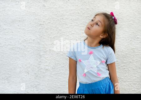 Ein denkendes Mädchen schaut nach oben und nach links auf einen leeren Raum für ein Design oder Logo oder Text. Das Kind träumt von etwas gegen eine leere weiße Wand. Stockfoto