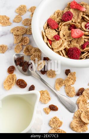Draufsicht auf Schüssel mit Getreide, roten Beeren, Löffel und Krug mit Milch, auf weißem Marmortisch, in vertikaler Richtung Stockfoto