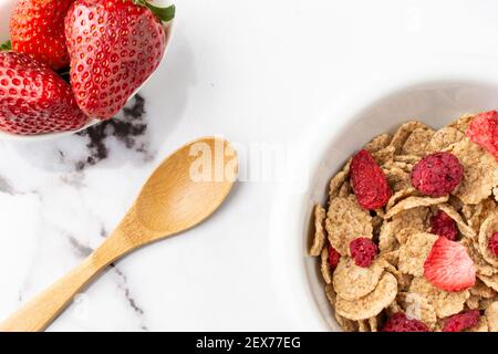 Luftaufnahme einer Schale mit Getreide, roten Beeren, Schale mit Erdbeeren und Holzlöffel, auf weißem Marmortisch, horizontal, mit Kopierplatz Stockfoto