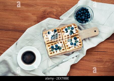 Waffeln mit Sahne und Heidelbeeren, eine Tasse Kaffee auf dem Tisch auf einer Stoffserviette. Frühstückskonzept. Draufsicht, flach liegend. Stockfoto