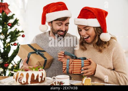 Aufgeregt Paar in weihnachtsmann Hüte geben Geschenke bei jedem Andere während wir Weihnachtsessen in gemütlichen Zimmer Stockfoto