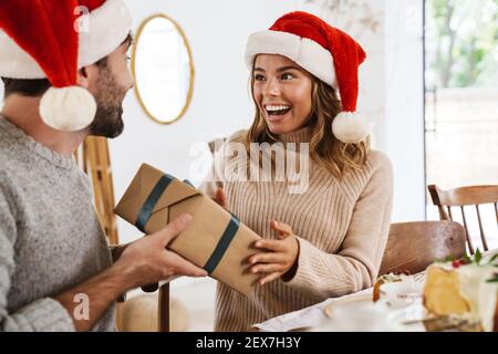 Glücklicher Mann in weihnachtsmann Hut geben Geschenkbox zu Seine Freundin beim Weihnachtsessen im gemütlichen Zimmer Stockfoto