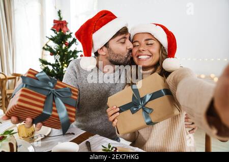 Nizza aufgeregt paar küssen und Selfie mit Geschenkboxen Während des Weihnachtsdinners in gemütlichem Zimmer Stockfoto