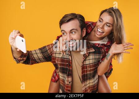 Aufgeregt Mann nimmt Selfie auf Handy, während Huckepack Reiten seine Freundin isoliert auf gelbem Hintergrund Stockfoto