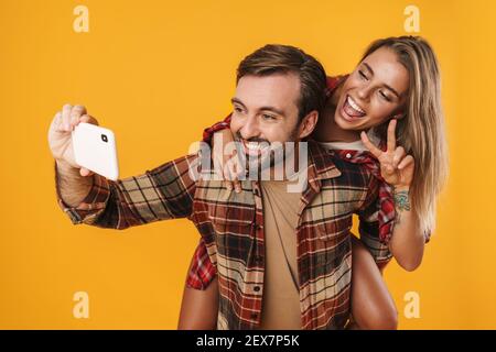 Aufgeregt Mann nimmt Selfie auf Handy, während Huckepack Reiten seine Freundin isoliert auf gelbem Hintergrund Stockfoto