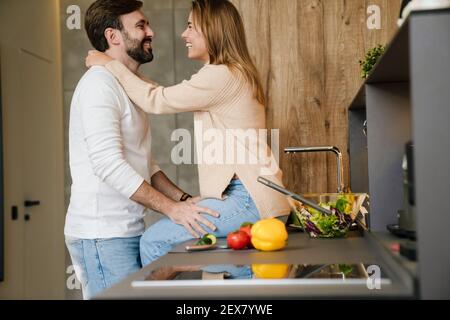 Lächelndes junges Paar kocht Salat zusammen in der Küche, umarmt Stockfoto