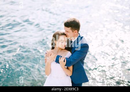 Der Bräutigam umarmt die Braut von hinten an den Schultern An der Küste Stockfoto