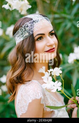 Die Braut steht bei einem blühenden weißen Oleander und hält Ein Zweig Oleander in ihren Händen Stockfoto