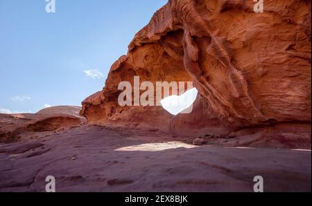 Kleiner Bogen oder kleine Felsenfensterformation in der Wadi Rum Wüste, helle Sonne scheint auf rotem Staub und Felsen, blauer Himmel darüber Stockfoto