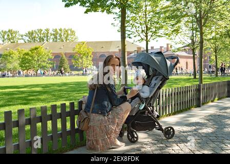 Porträt eines entzückenden Kleinkindes und seiner schönen Mutter, die in einem Kinderwagen sitzt und die Kamera auf dem Hintergrund des öffentlichen Parks anschaut. Stockfoto