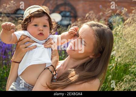 Portrait einer jungen schönen Mutter mit einem launischen und frechen kleinen Jungen in den Armen an einem Sommertag im Freien. Stockfoto