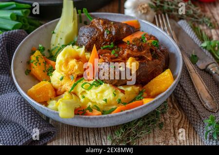 Ossobuco, geschmorte Rinderschenkel mit Kartoffelpüree und Fenchel, Karotten, Orangen Gemüse serviert mit köstlicher Soße auf einem Teller auf rustikalem Tisch Stockfoto