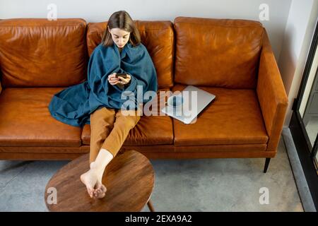 Hübsche Frau mit Smartphone bedeckt mit einem blauen karierten Sitz auf dem braunen Sofa zu Hause. Lifestyle und Wohnlichkeit Konzept. Stockfoto