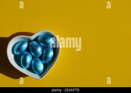 Weiße herzförmige Schale mit blauer Folie verpackt Schokolade Ostern Eier isoliert auf hellgelbem Hintergrund mit Kopierraum Stockfoto