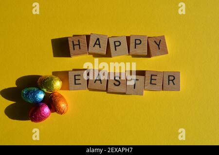 Frohe Ostern, Wörter in hölzernen Alphabet Buchstaben und Folie verpackt Schokoladeneier isoliert auf hellem gelben Hintergrund Stockfoto