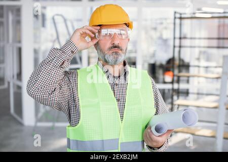 Hoch qualifizierte bärtige Mann in Brille und Helm posiert mit Blaupausen Stockfoto