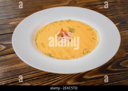 Kürbiscremesuppe garniert mit Speckscheiben und Rettich auf einem weißen Teller auf einem Holztisch. Stockfoto