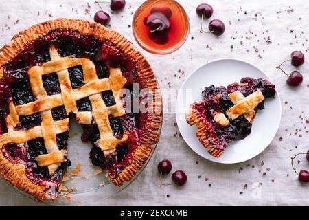 Flatlay Stillleben einer gebackenen Kirschkuchen mit einer Scheibe herausgenommen und sitzt auf einem Teller in der Nähe eines Cocktails. Stockfoto