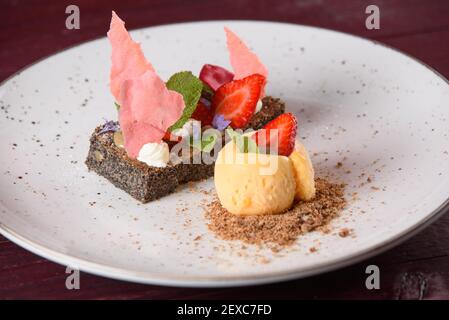 Poppyseed Dessert mit Eis und Keksbröseln, garniert mit Erdbeer-Schokolade, Erdbeeren, Minze, Sahne und Blumen auf einem weißen Teller. Stockfoto