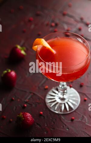 Erdbeer Gin gepflegter Cocktail in einem Vintage Coupé Glas, umgeben von Erdbeeren und rosa Pfefferkörnern auf einer roten Oberfläche. Stockfoto