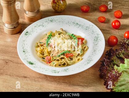 Nudeln mit Käse und Tomaten in einer weißen Sauce mit Kräutern und Kirschtomaten auf einem weißen Teller auf einem Holztisch. Stockfoto