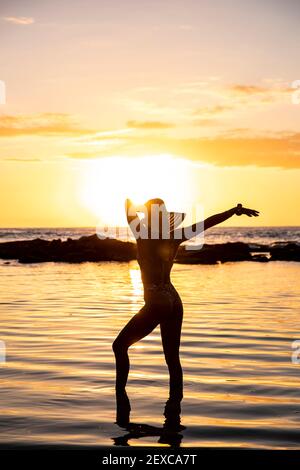 Frau posiert in seichtem Wasser bei Sonnenuntergang trägt Hut und Bikini Stockfoto