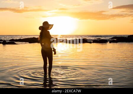 Weibchen steht im seichten Wasser des Ozeans mit Hut Bei Sonnenuntergang Stockfoto