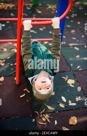 Niedliches Kind, das im Herbst kopfüber auf Affenstangen hängt Spielplatz