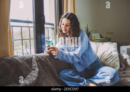 Junge Frau, die das Handy auf dem Sofa in der Nähe betrachtet Das Fenster Stockfoto